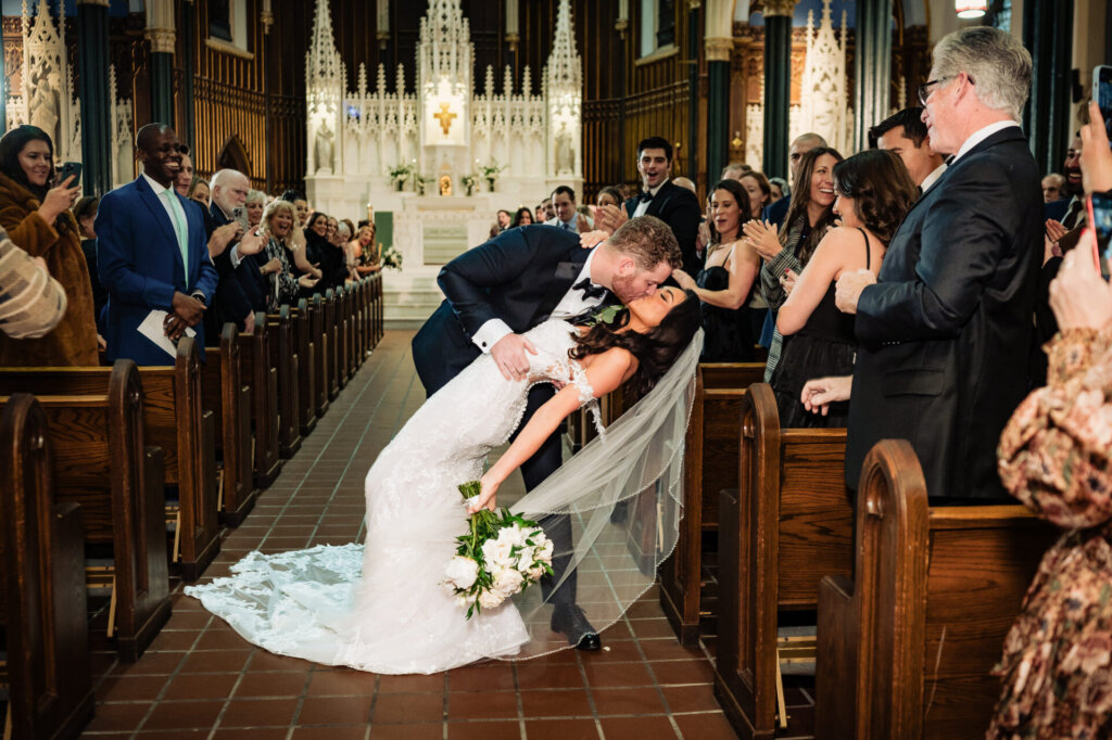 Groom dipping the bride for a romantic kiss in the church aisle, her bouquet resting on the floor as guests applaud around them.