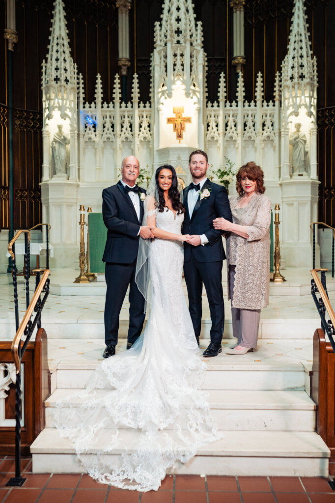 Bride and groom posing with the bride’s parents at the church altar, her lace train cascading down the steps and the ornate backdrop enhancing the moment