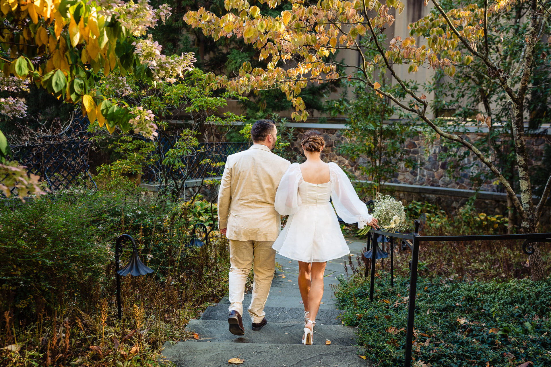 A couple walking hand in hand on a garden pathway, the man wearing a beige suit and the woman in a white dress with flowing sleeves, surrounded by autumn foliage