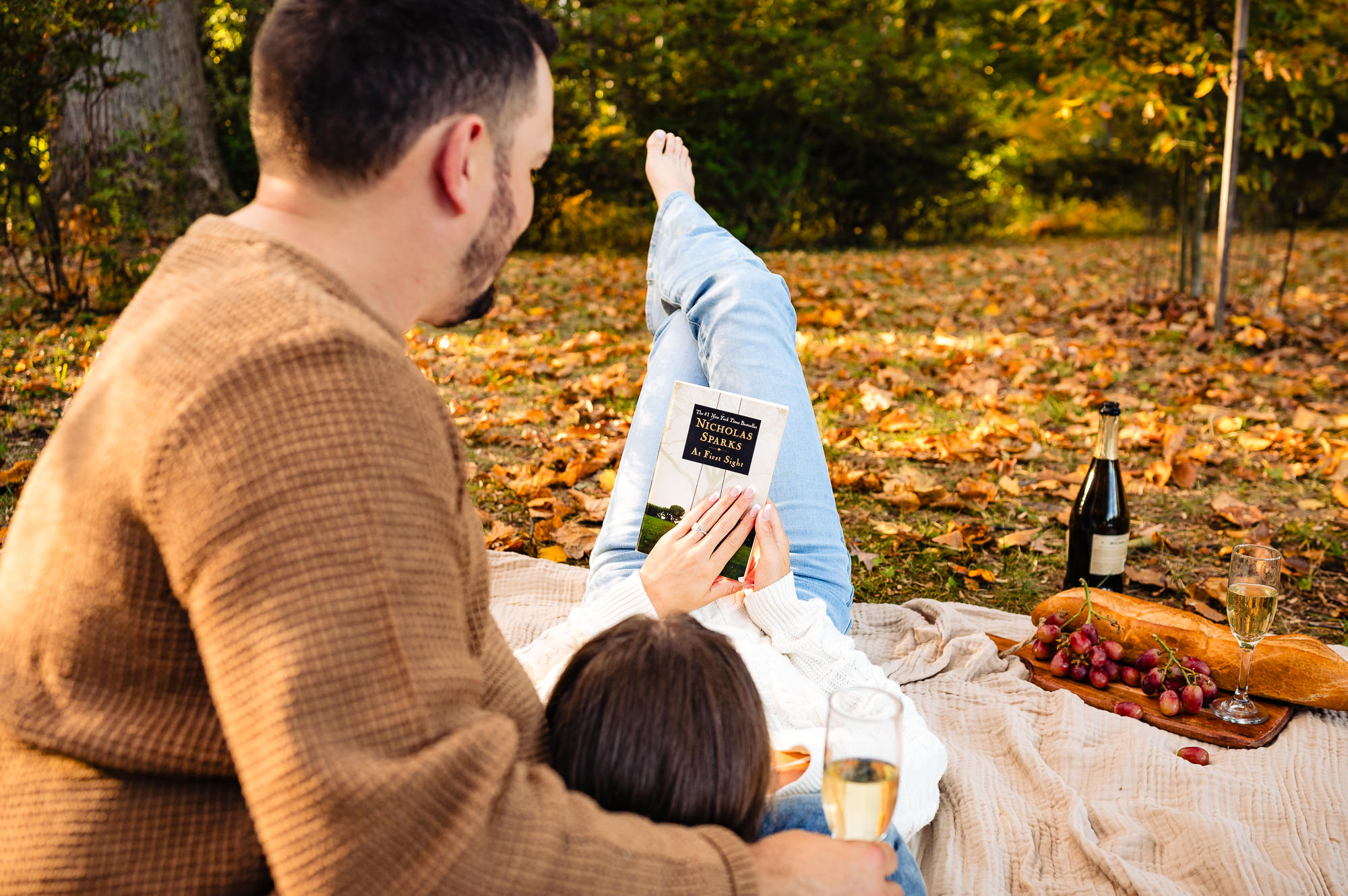 A creative perspective showing the couple lying on the blanket, holding a book and wine glasses, with autumn colors in the background.