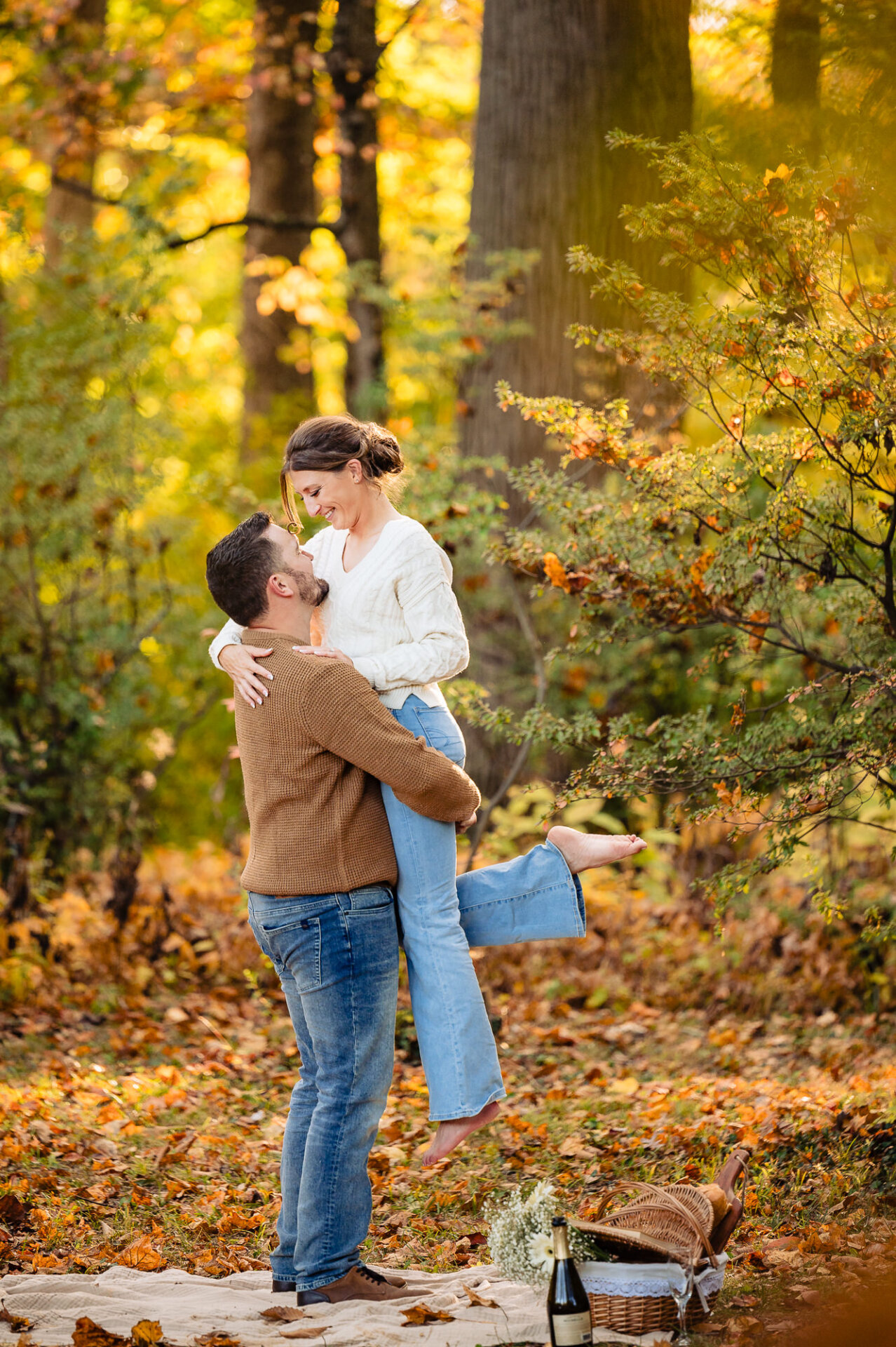 A couple sharing a joyful embrace as the man lifts the woman in a vibrant autumn forest, surrounded by golden leaves. A picnic setup with a basket, wine, and flowers adds warmth to the romantic scene.
