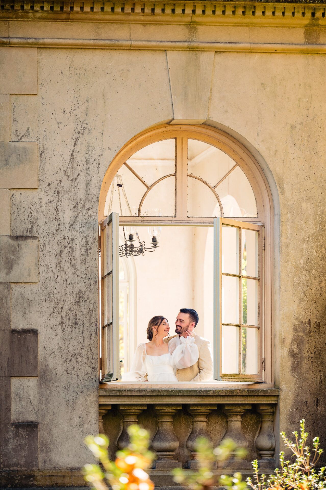 A couple smiling warmly at each other, standing in a beautifully arched window with delicate architectural details.