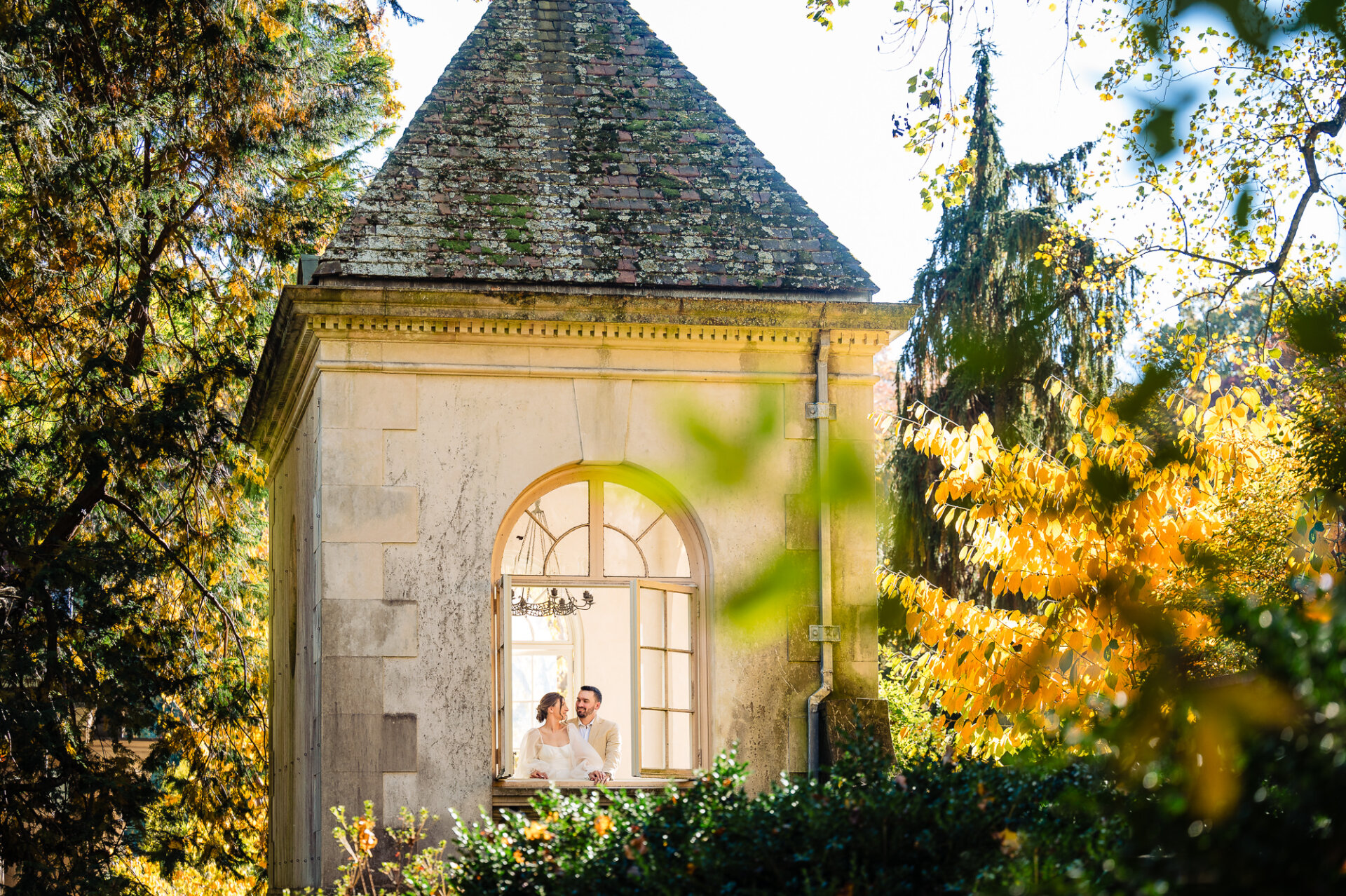 A romantic scene of a couple standing inside a rustic tower with an arched window, surrounded by vibrant autumn foliage and a tranquil garden setting