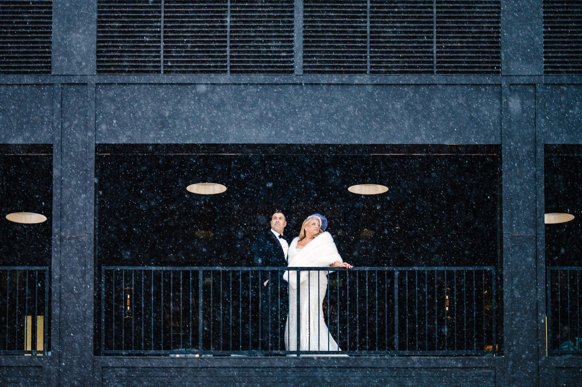 Bride and groom dancing on the balcony of a modern black building during snowfall
