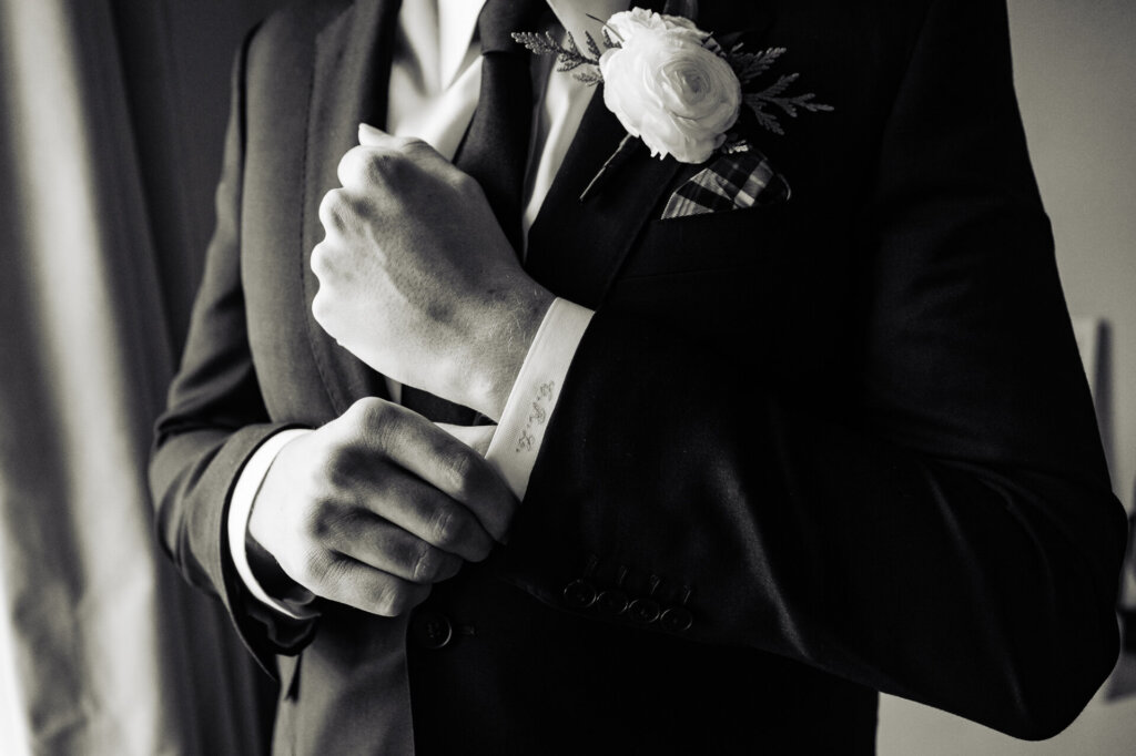 A detail shot of the groom's hands as he fastens his cufflinks, wearing a black suit with a white boutonniere.