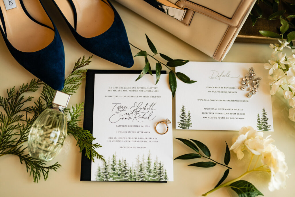 A nature-inspired wedding invitation suite displayed alongside blue heels, perfume, and fresh greenery.
