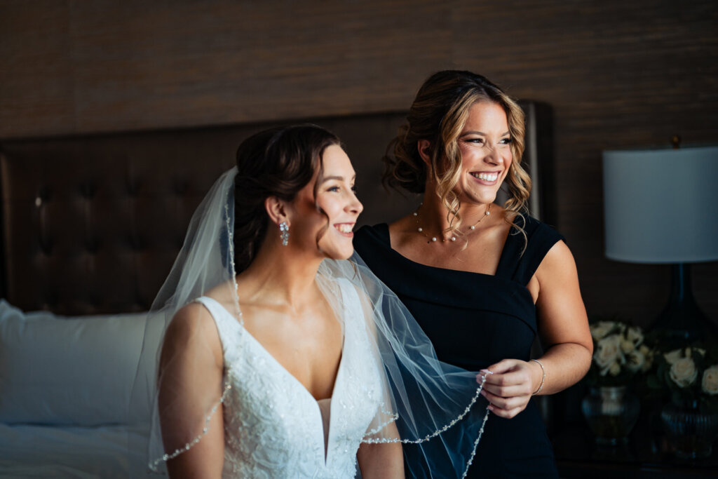 The mother of the bride helps adjust her daughter’s gown, both smiling in a warmly lit room.