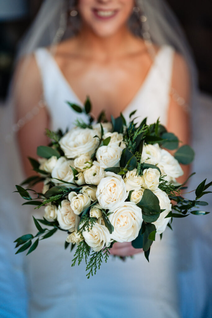 A close-up image of the bride’s bouquet filled with fresh white roses and greenery, with a blurred smile in the background.
