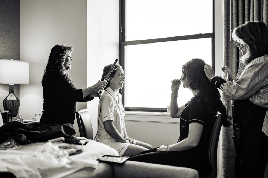 A black-and-white candid moment of the bridal party helping the bride get ready near a bright window.