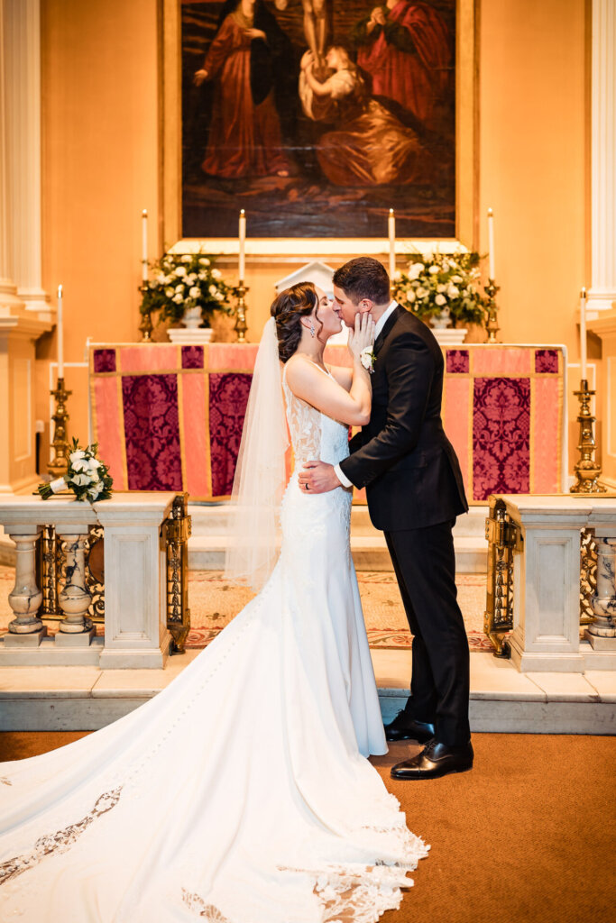 A romantic moment as the newlyweds embrace in front of a grand altar, surrounded by floral arrangements and candlelight.