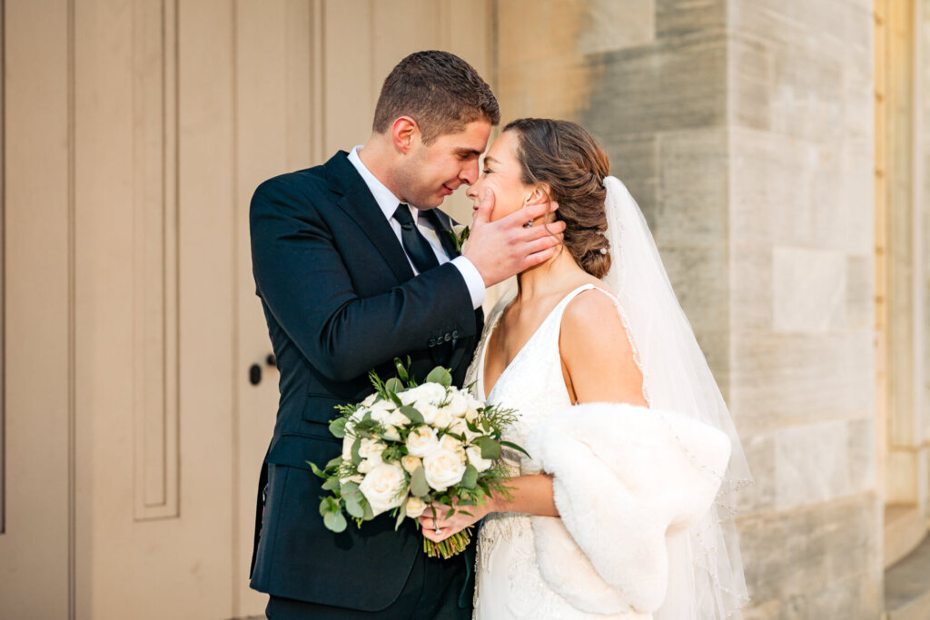 Couple leaning in each other captured in front of a grand, textured wall, with the bride’s bouquet in hand.