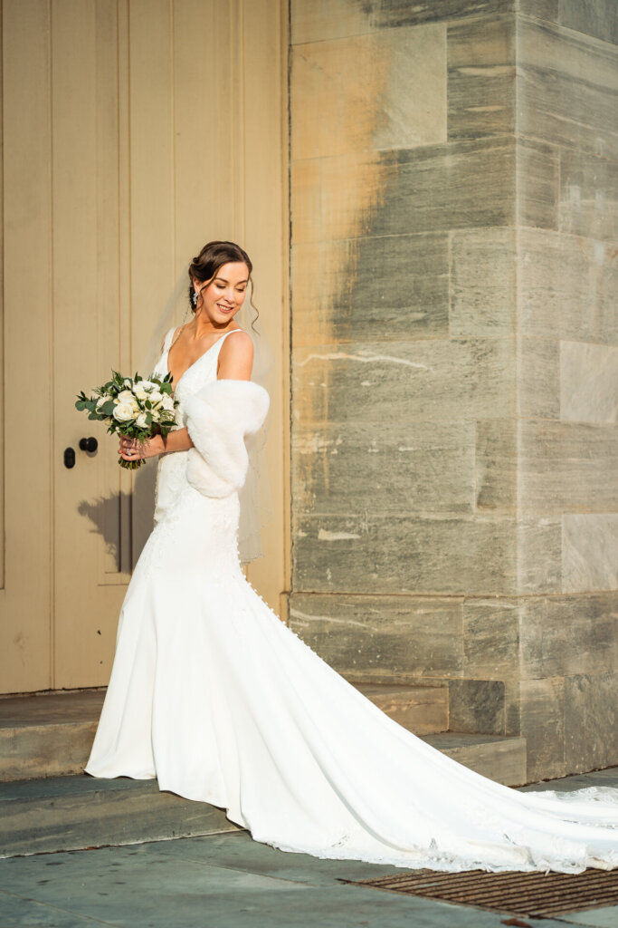 A full-length portrait of the bride in her lace gown, wrapped in a soft fur stole, smiling with grace.