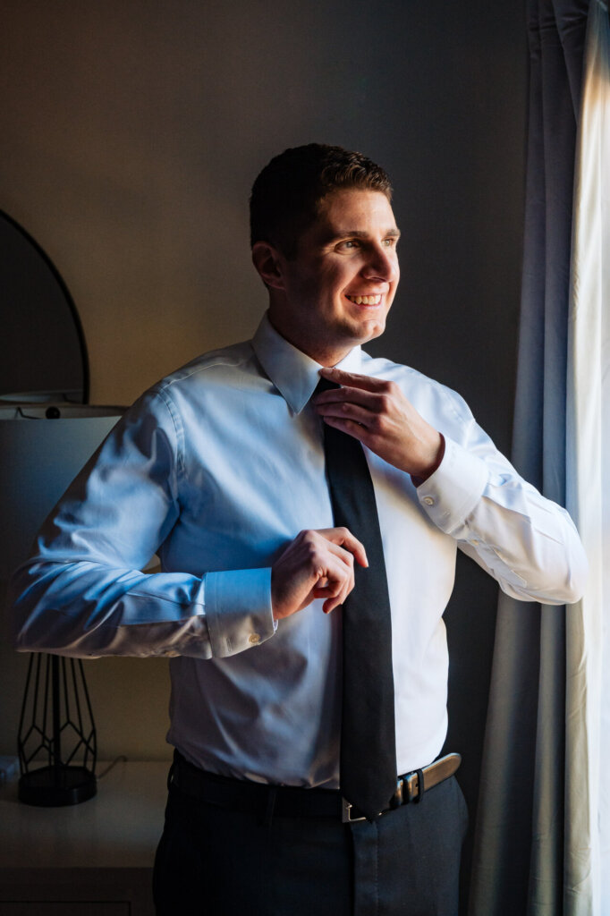 A smiling groom stands by a window, fixing his tie as natural light highlights his face.
