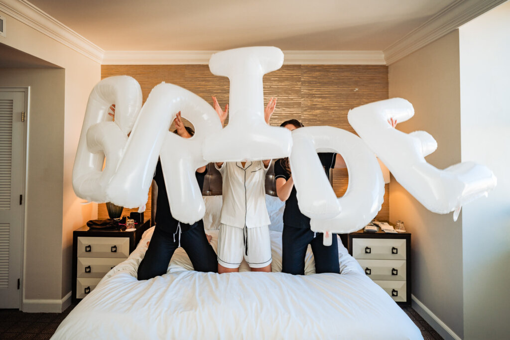 The bridal party joyfully tosses white balloons in a hotel room, laughing together before the ceremony