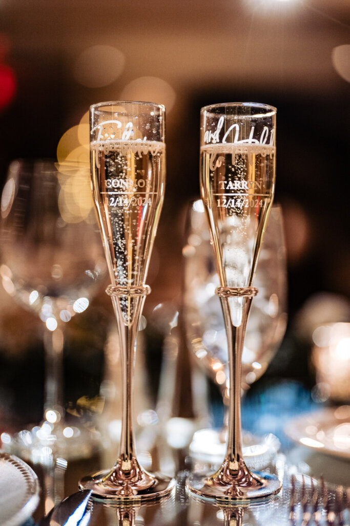 Two crystal champagne glasses engraved with the wedding date, filled with bubbly, set against a softly lit reception backdrop.