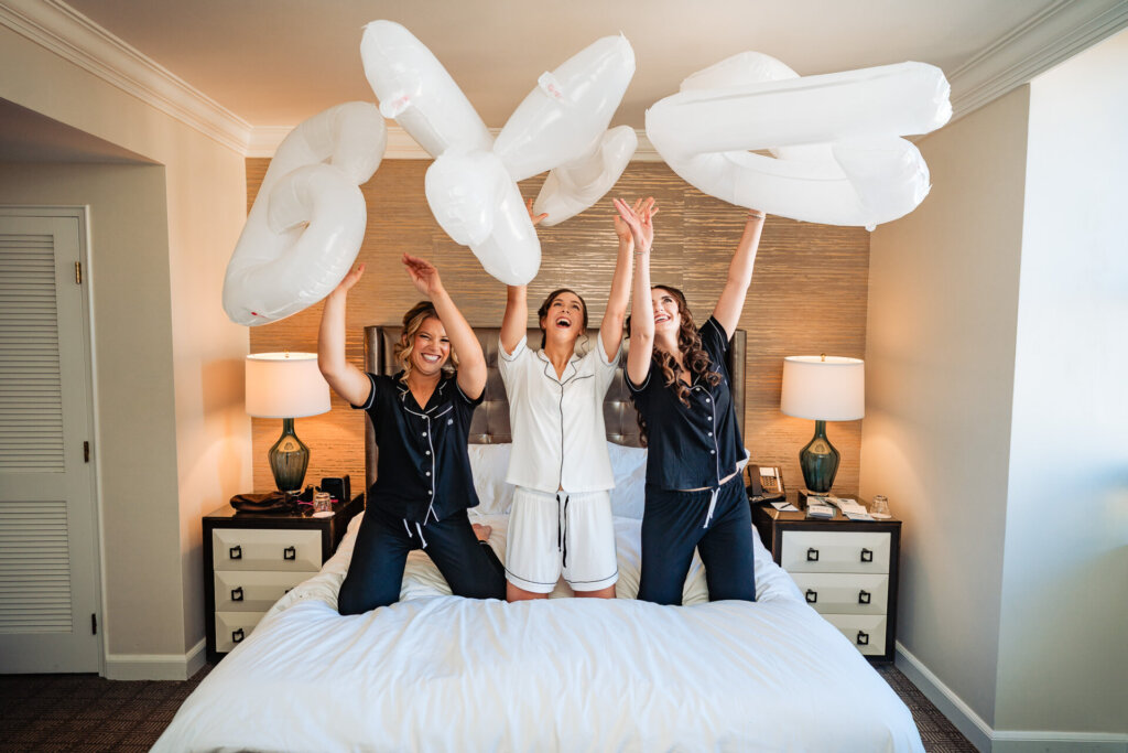 The bridal party joyfully tosses white balloons in a hotel room, laughing together before the ceremony.